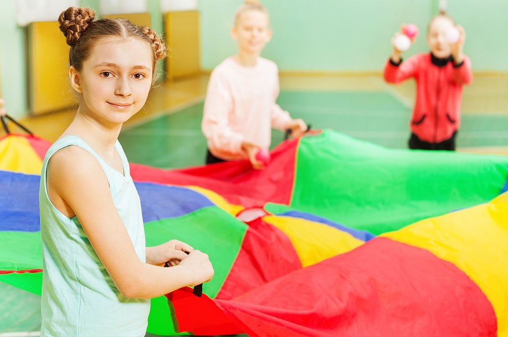 girl with braids smiling,, holding a parachute with friends in a gymnasium