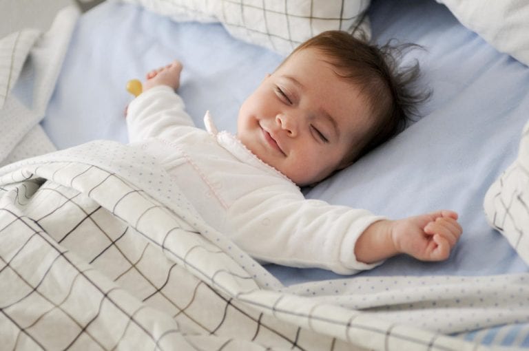 smiling baby sleeping with arms outstretched face up in bed
